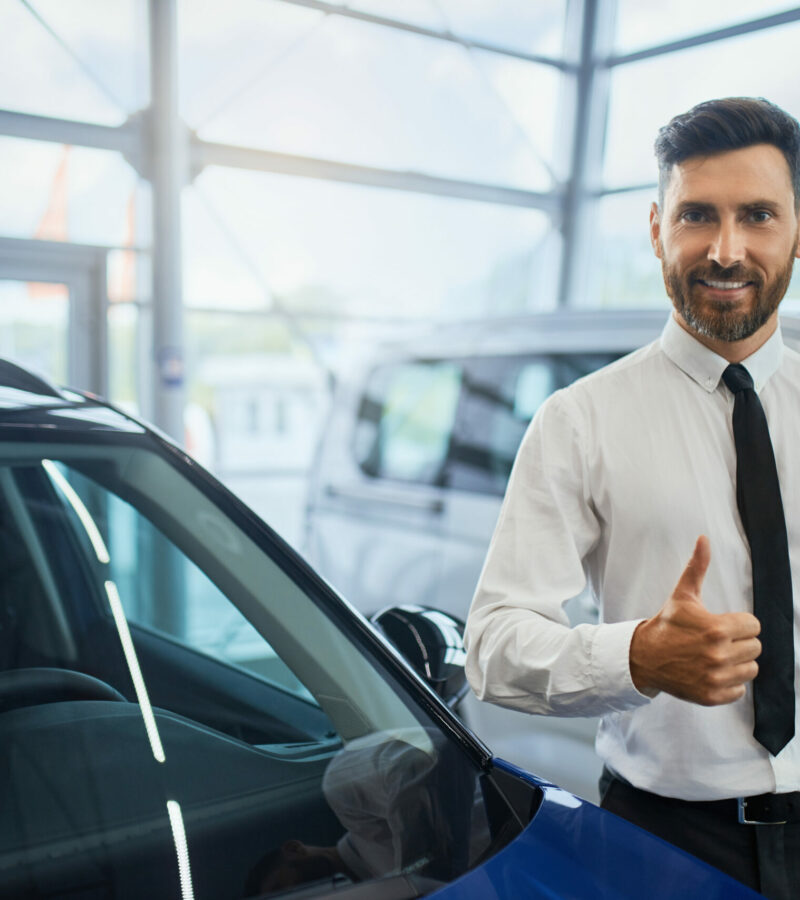 Portrait of handsome bearded man in formal clothes showing thumb up and smiling on camera while standing at auto salon. Male customer making expensive purchase.